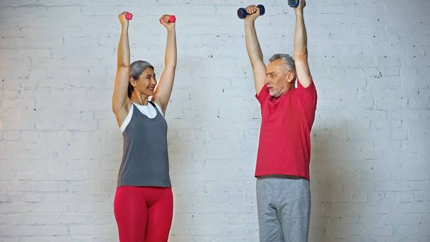 smiling asian woman looking at senior husband while training with dumbbells - Photo, Image