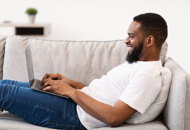 Czarny wolny strzelec pracuje na laptopie leżąc na kanapie w domu - Zdjęcie, obraz