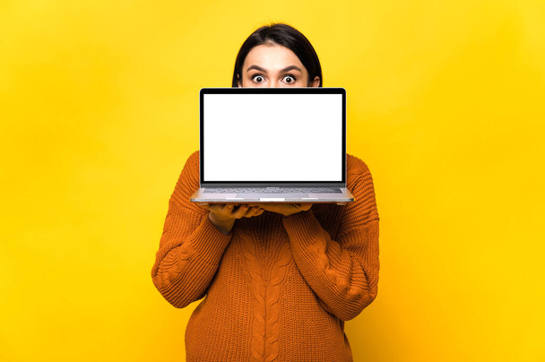 Sorprendido sorprendido mujer joven caucásica, escondido detrás de la pantalla blanca en blanco de la computadora portátil, de pie sobre un fondo naranja aislado. Copiar espacio. - Foto, Imagen