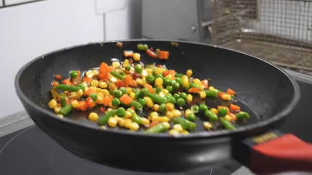 Male arm of cook holding skillet and tossing up mixed vegetables at kitchen restaurant. Chef sauteing corn, peas, asparagus, pepper and carrot on olive oil at frying pan. Concept of cooking food - Footage, Video