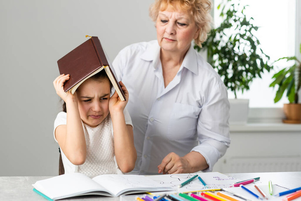 Woman tutor or foster parent mum helping cute caucasian school child girl doing homework sitting at table. Diverse nanny and kid learning writing in notebook studying at home. - Photo, Image