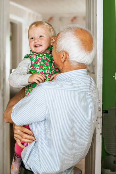 Siwy dziadek z blond włosami w ramionach. Głowa dziadka i wnuczki jest blisko. Zbliżenie zdjęcie śmieszne dwie osoby stary dziadek mała wnuczka - Zdjęcie, obraz