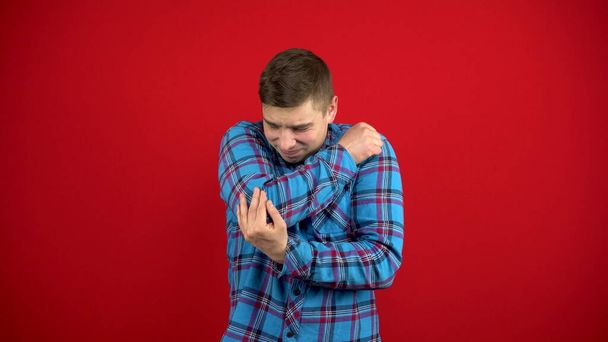 A young man has a sore elbow and holds it in his hand. Shooting on a red background. - Photo, Image