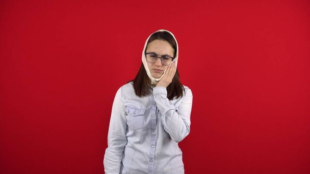A young woman with glasses has a rewound head due to a pain in a tooth. The girl touches her cheek with her hand. Shooting on a red background. - Photo, Image