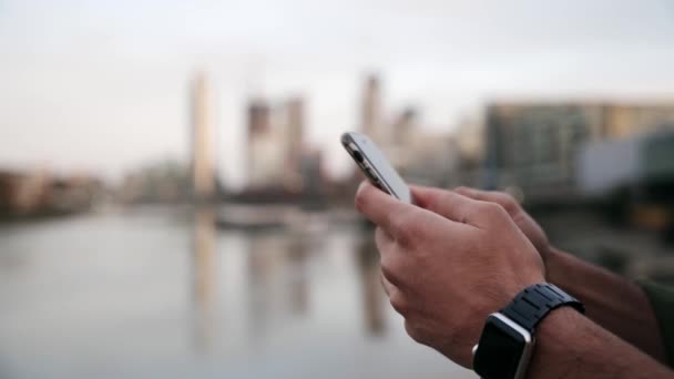 Lähikuva sekoitettu rotu mies kirjoittamalla älypuhelimeen seisoo kaupungin näkymät järvelle - Materiaali, video