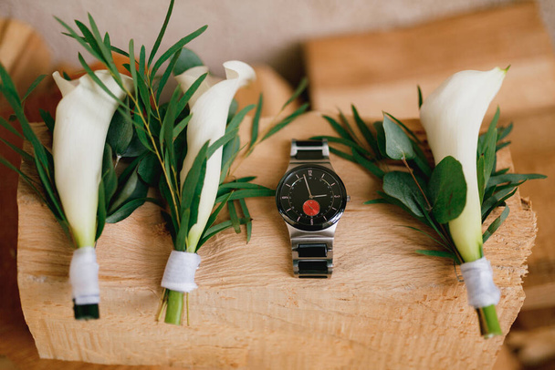 Close-up of a black mens watch on a wooden texture with boutonnieres with white calla lilies. - Photo, Image