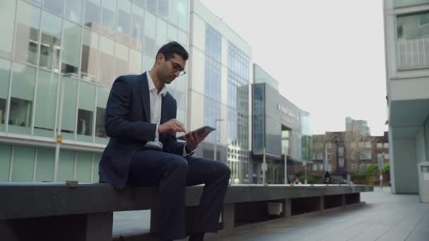 Бизнесмен смешанной расы печатает на цифровом планшете, сидя за пределами офисного здания - Кадры, видео