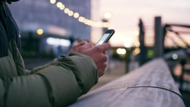 Lähikuva sekoitettu rotu uros nuori aikuinen tekstiviesti älypuhelimeen nojaten sillan kaiteeseen kylmässä kaupungissa illalla  - Materiaali, video