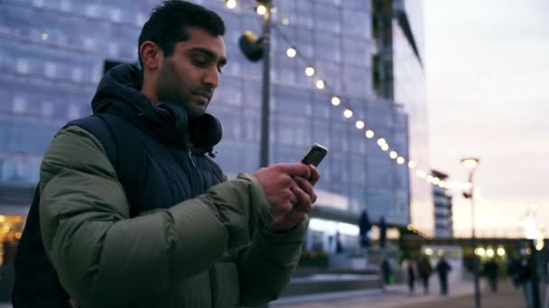 Sekarotuinen uros nuori aikuinen hymyilee ja nauraa chattailuun tyttöystävä älypuhelin seisoo kylmässä kaupungissa ilta  - Materiaali, video