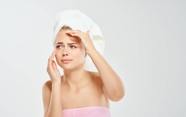 γυναίκα με λευκή πετσέτα στο κεφάλι αγγίζει το πρόσωπό της με τα χέρια της σε ελαφρύ φόντο - Φωτογραφία, εικόνα