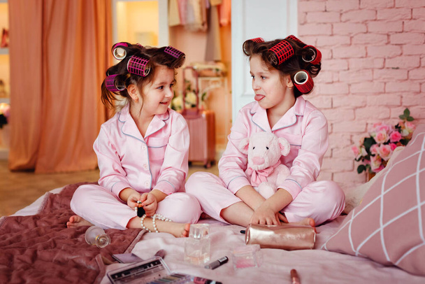 Τα κοριτσάκια με τις ροζ πιτζάμες βάφονται ενώ κάθονται σε ένα δωμάτιο. Αδελφές με μπικουτί παίζουν με αξεσουάρ μακιγιάζ στο παιδικό δωμάτιο. Τα παιδιά κάθονται στο κρεβάτι με το μακιγιάζ της μητέρας τους. - Φωτογραφία, εικόνα
