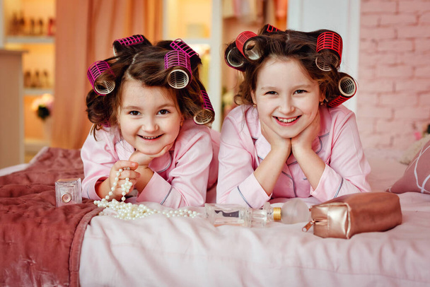 Holčičky v růžovém pyžamu se líčí, když sedí v pokoji. Sestry s natáčkami hrát s make-up příslušenství v dětském pokoji. Děti sedí na posteli s matčiným make-upem - Fotografie, Obrázek