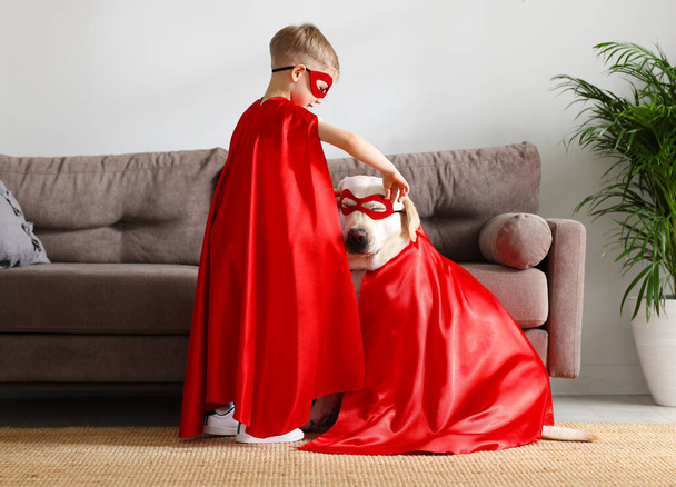 Vista lateral de cuerpo completo de niño pequeño y lindo perro Labrador retriever vestido con capas de superhéroes rojos y gafas jugando juntos en casa - Foto, imagen