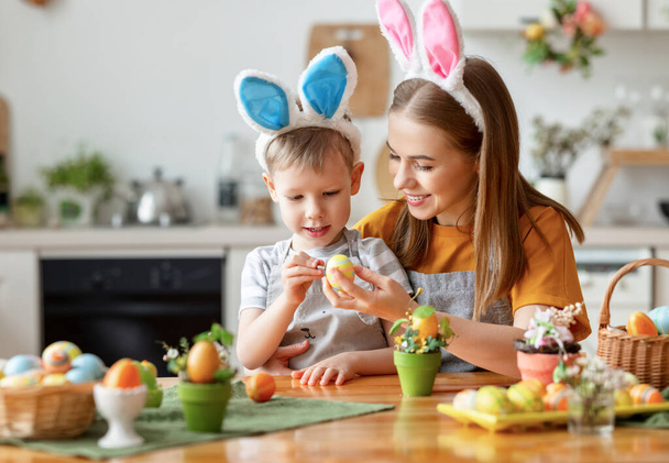 Neşeli genç bir anne ve sevimli bir oğul komik tavşan kulaklıkları takıp mutfakta paskalya yumurtalarını boyuyorlar.   - Fotoğraf, Görsel
