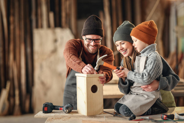 Menino focado usando martelo ao fazer casa de pássaro de madeira com a mãe e o pai na oficina de marcenaria profissional - Foto, Imagem