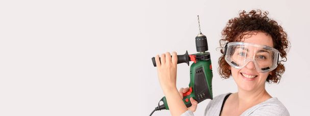 Caucasian woman with curly hair holds a drill in her hands, ready to make home improvements. She wears a gray shirt. White background. Natural light. Background with copy space. - Photo, Image