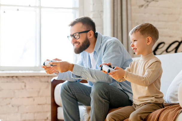 Happy family: bearded man and delighted boy using gamepads to play videogame together while chilling on settee in weekend at home - Photo, Image