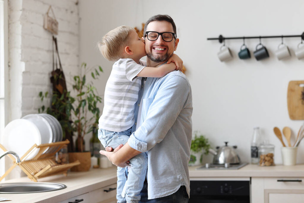Visão lateral conteúdo jovem pai segurando bonito pequeno filho a mãos e recebendo doce bochecha beijo enquanto está de pé na cozinha moderna - Foto, Imagem