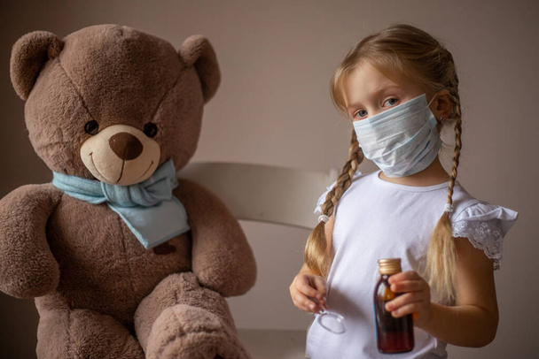 Una niña pequeña en una máscara trata a un oso de juguete grande en una bufanda, le da medicina, medicina. juega en el médico de los niños. Concepto de salud, vacunación de niños, pediatra. Enfoque selectivo - Foto, Imagen