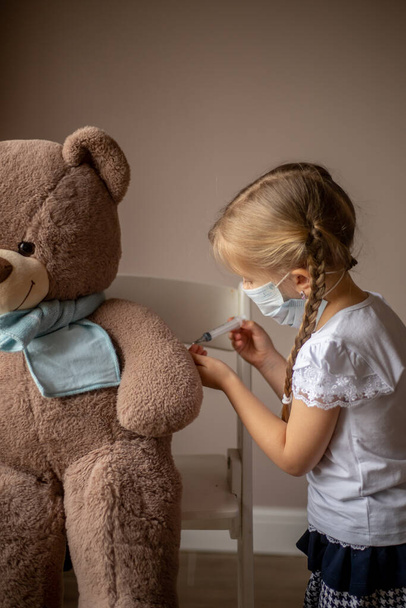 A little girl with pigtails in a mask makes an injection, an inoculation to a big toy bear in a scarf, treats, plays a child's doctor. Health concept, vaccination of children, pediatrician - Photo, Image