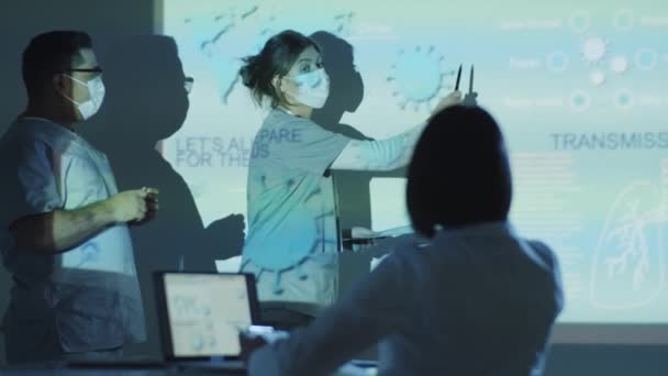 Mężczyzna i kobieta naukowcy w mundurach medycznych i maskach ochronnych omawiający prezentację o wirusie covid-19 z kolegą w ciemnym laboratorium - Materiał filmowy, wideo
