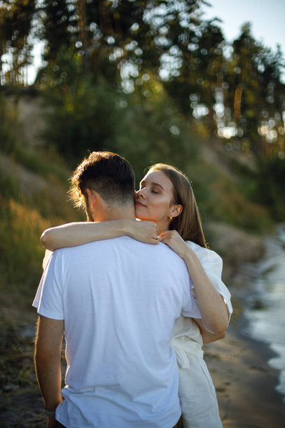 muž s ženou v bílém se něžně objímají na březích řeky, jezera nebo moře. milující pár objímání na pláži při západu slunce - Fotografie, Obrázek