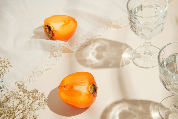 Moitiés de fruits kaki et deux verres d'eau sur fond pastel avec chiffon blanc et lumière du soleil. Boissons d'été et concept de rafraîchissement. Composition de style minimal - Photo, image