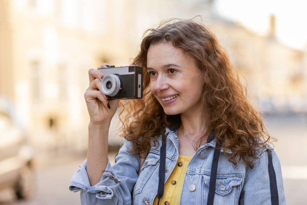 Cheerful curly hair woman tourist with retro camera walking along city street on bright sunny day. Travel concept - Fotografie, Obrázek