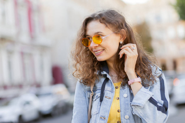 Atrakcyjne, kręcone włosy w średnim wieku kobieta z żółtymi okularami przeciwsłonecznymi w stylowym drelichu ubrania w mieście w jasny słoneczny dzień - Zdjęcie, obraz