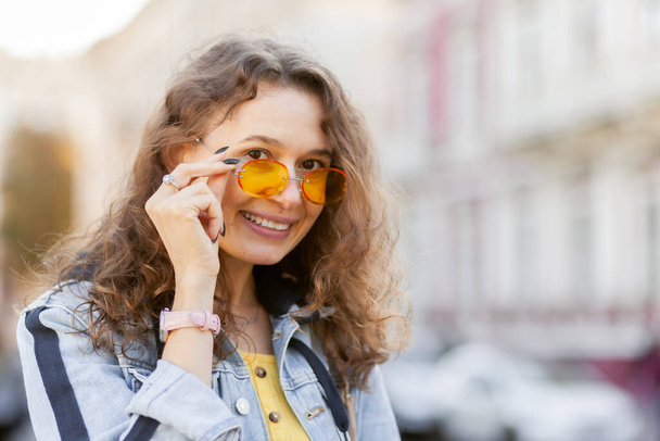 Среднего возраста привлекательная кудрявая женщина с желтыми солнцезащитными очками в стильной джинсовой одежде в городе в яркий солнечный день - Фото, изображение