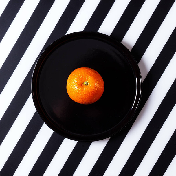 Naranja sobre plato negro sobre fondo blanco y negro rayado. Diseño plano, vista superior. - Foto, imagen