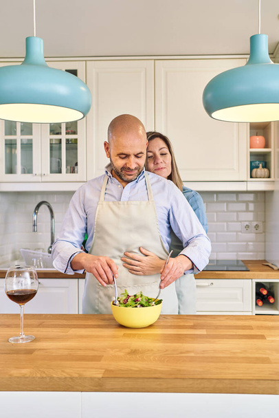 Een man die salade maakt terwijl zijn vrouw hem van achteren knuffelt. Koken als koppel - Foto, afbeelding