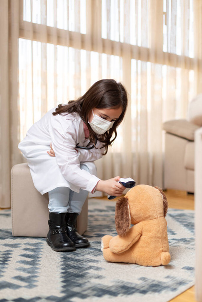 Klein kind in doktersuniform en medisch masker neemt de temperatuur van een teddybeer op met een infrarood thermometer. Ze is thuis. Ruimte voor tekst. - Foto, afbeelding