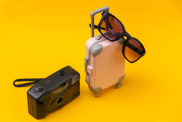 Koncepcja podróży. Mini różowa plastikowa walizka podróżna z kamerą, okulary przeciwsłoneczne na żółtym tle. Minimalny styl - Zdjęcie, obraz
