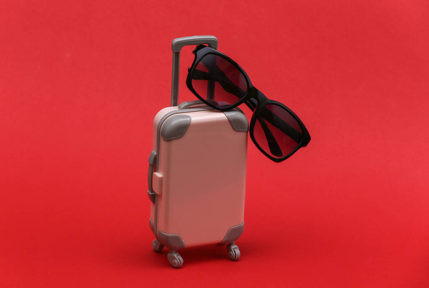 Koncepcja podróży lub podróży. Mini plastikowa walizka podróżna w okularach przeciwsłonecznych na czerwonym tle. Minimalny styl - Zdjęcie, obraz