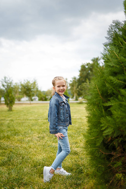 Malá usměvavá dívka v džínové bundě pózující venku v jarním parku na zeleném pozadí keře. Happy optimistic child, carefree childhood concept - Fotografie, Obrázek