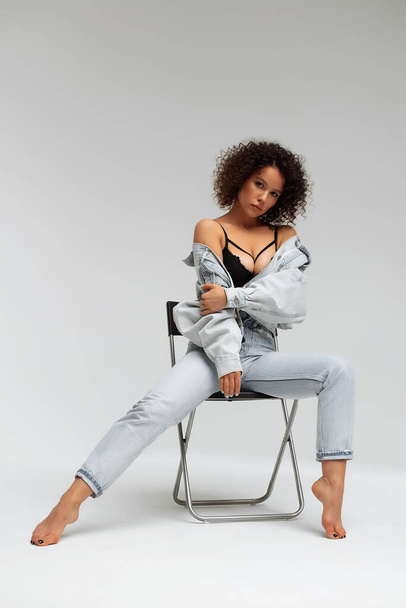 Полное тело уверенной молодой босиком модели с кудрявой прической в модном джинсовом пиджаке и джинсах, сидящих на стуле в белой студии - Фото, изображение