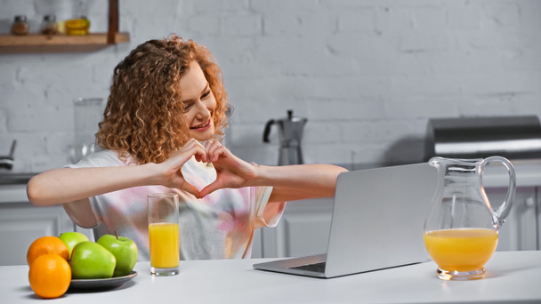 σγουρά νεαρή γυναίκα κοιτάζοντας το laptop, ενώ δείχνει την καρδιά σημάδι με τα χέρια κατά τη διάρκεια της κλήσης βίντεο - Φωτογραφία, εικόνα