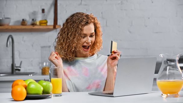 excité jeune femme tenant carte de crédit tout en faisant des achats en ligne dans la cuisine  - Photo, image