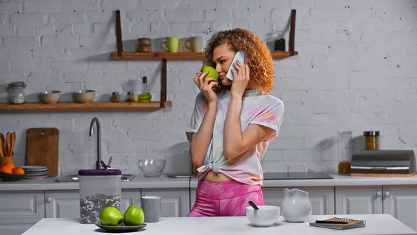 göndör nő beszél okostelefon és szaga alma közelében kukoricapehely konténerben az asztalon - Fotó, kép