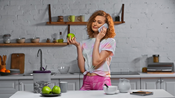 kudrnatá mladá žena mluví na smartphone a drží jablko v blízkosti kukuřičné vločky v kontejneru na stole - Fotografie, Obrázek