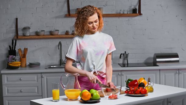 curly young woman cutting fresh tomato in kitchen  - Φωτογραφία, εικόνα