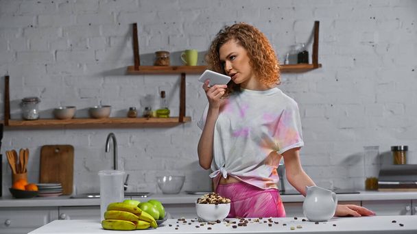 mulher encaracolado gravação mensagem de voz perto de flocos de milho e frutas na mesa da cozinha - Foto, Imagem