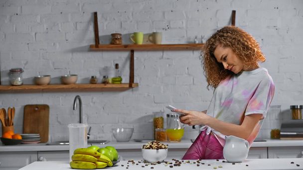 femme bouclée prenant des photos de flocons de maïs et de fruits sur la table de cuisine - Photo, image
