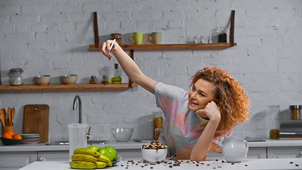 кудрявая молодая женщина делает селфи возле кукурузных хлопьев и фруктов на кухонном столе - Фото, изображение