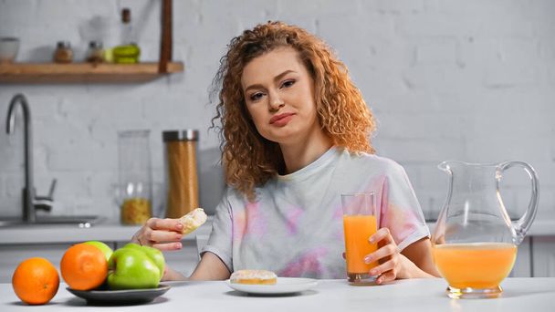 smiling woman holding doughnut and glass of orange juice in kitchen  - Foto, afbeelding