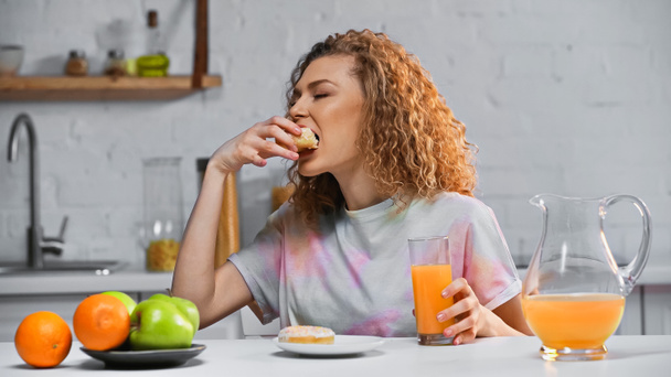 curly woman eating sweet doughnut and holding glass of orange juice in kitchen  - Photo, Image