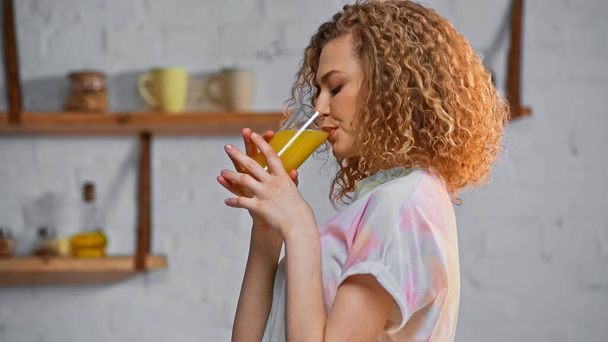 πλαϊνή άποψη του σγουρά γυναίκα πίνοντας χυμό πορτοκαλιού στην κουζίνα  - Φωτογραφία, εικόνα