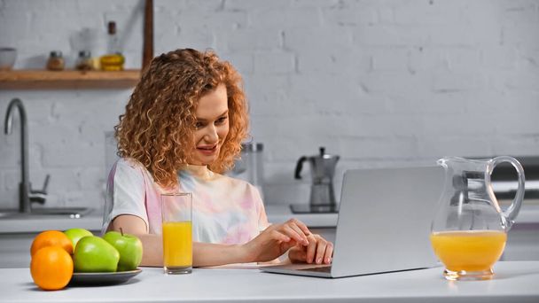 femme joyeuse regardant ordinateur portable près des fruits et un verre de jus d'orange sur la table - Photo, image