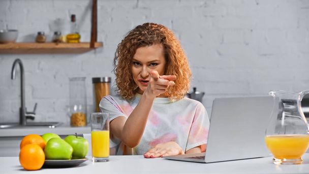 Kıvırcık kadın, dizüstü bilgisayarın yanındaki kameraya ve masadaki taze meyvelere bakarken parmağıyla işaret ediyor. - Fotoğraf, Görsel